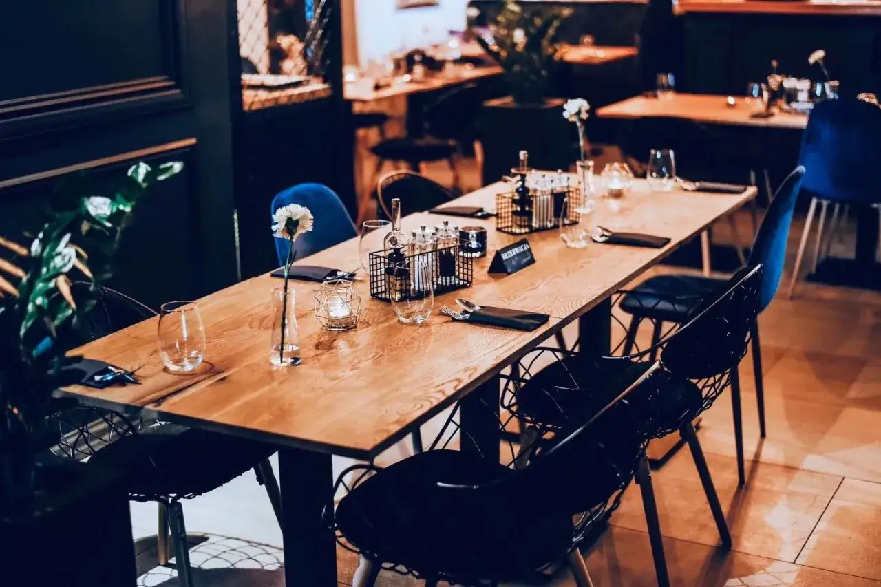 Elegancko nakryty stół w restauracji, gotowy na gości. Na drewnianym blacie kieliszki, świeca, kwiat i czarne serwetki. W tle widać inne stoliki i niebieskie krzesła. Idealne miejsce na romantyczną kolację lub spotkanie biznesowe, gdzie czeka **piernic...