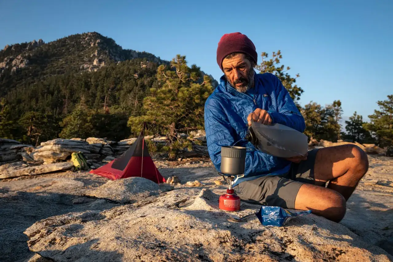 Vandrare förbereder mat vid sitt msr kök på en klippig bergssluttning med tält och skog i bakgrunden.
