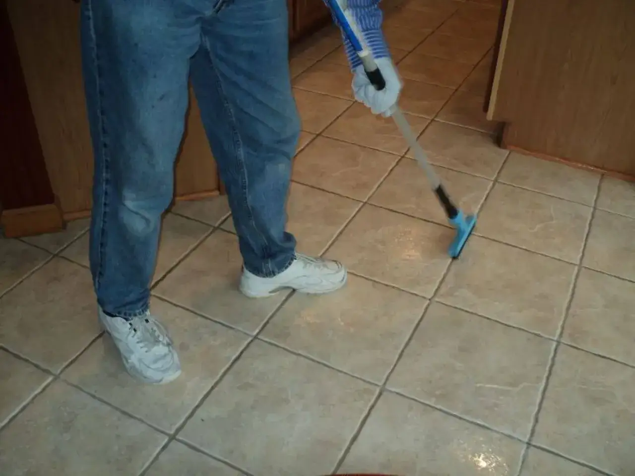 Persona con jeans e scarpe bianche pulisce le fughe del pavimento in piastrelle con uno strumento.