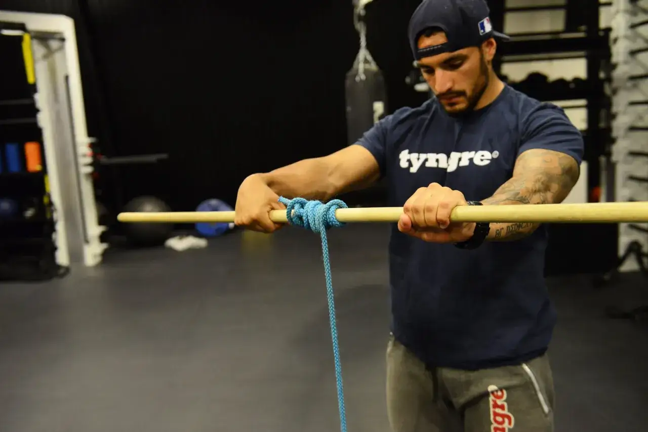 Man med keps och tatueringar stretcha armar medan han knyter ett rep runt en träpinne i ett gym.