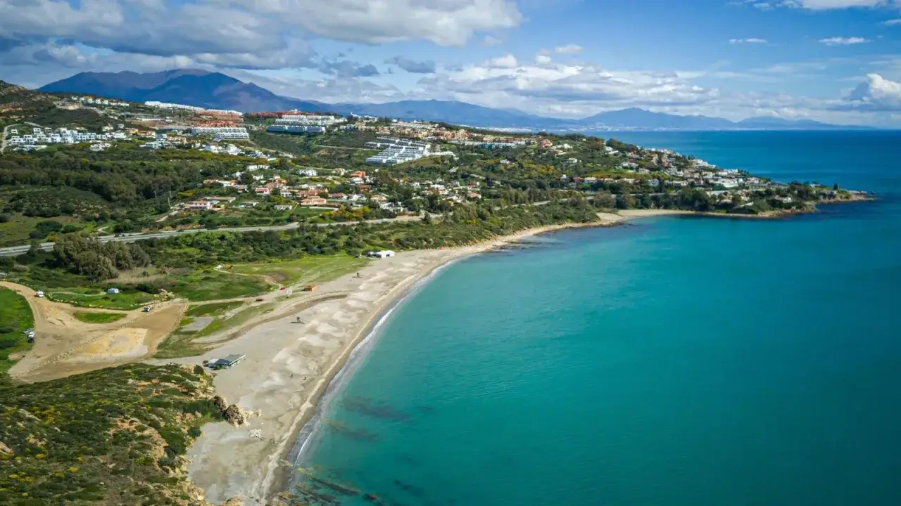 Vista aérea de una **playa** con aguas turquesas y colinas verdes cubiertas de casas. Un lugar perfecto para lucir tu **bañador** y disfrutar del sol.
