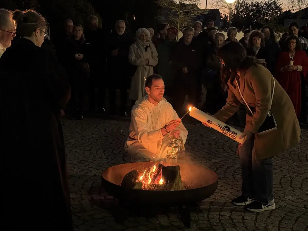 Menschen versammeln sich um ein Osterfeuer, eine alte Tradition. Eine Frau entz&uuml;ndet eine Osterkerze, w&auml;hrend ein Mann daneben sitzt.