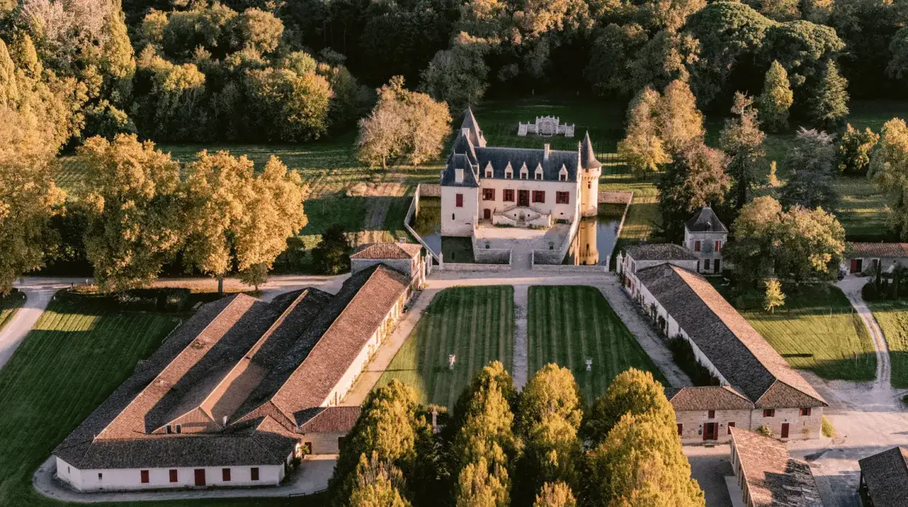 Vue a&eacute;rienne du **Ch&acirc;teau Olivier** entour&eacute; de douves, avec ses d&eacute;pendances et ses jardins &agrave; la fran&ccedil;aise baign&eacute;s par la lumi&egrave;re dor&eacute;e du soleil couchant.