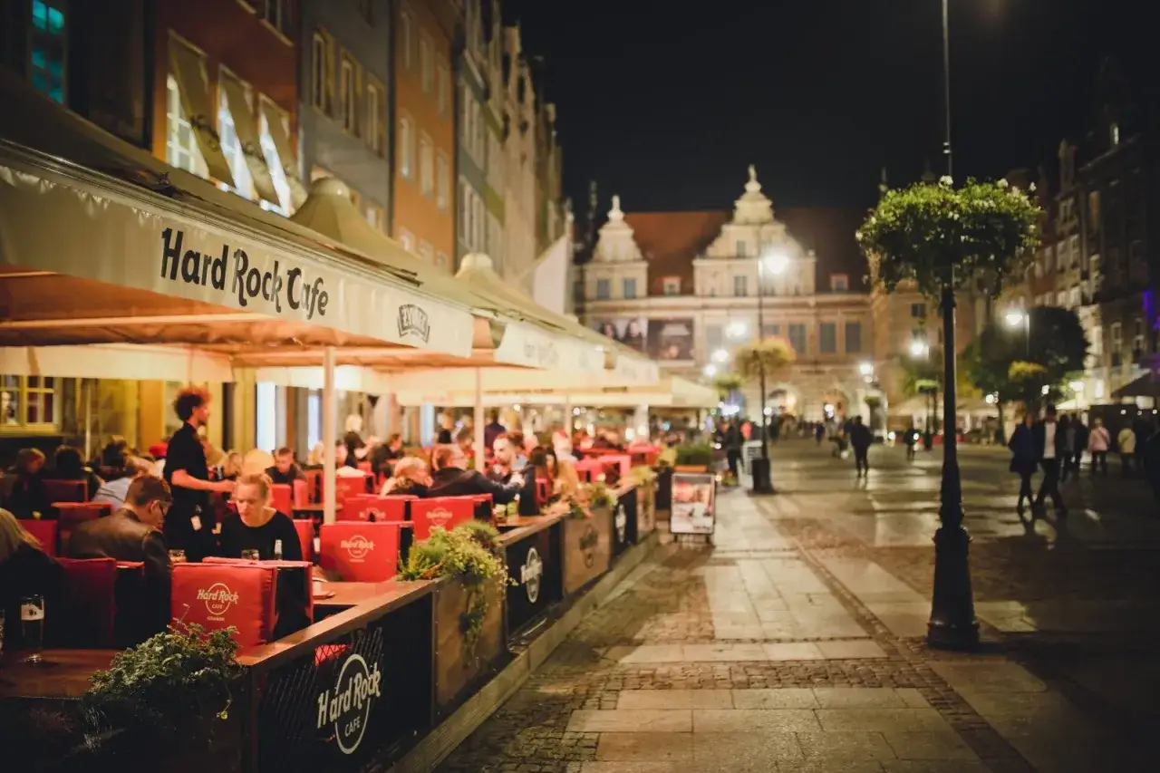 Wieczorny gwar przy stolikach Hard Rock Cafe w Gdańsku. Ludzie siedzą pod markizami, delektując się posiłkami i atmosferą miasta.