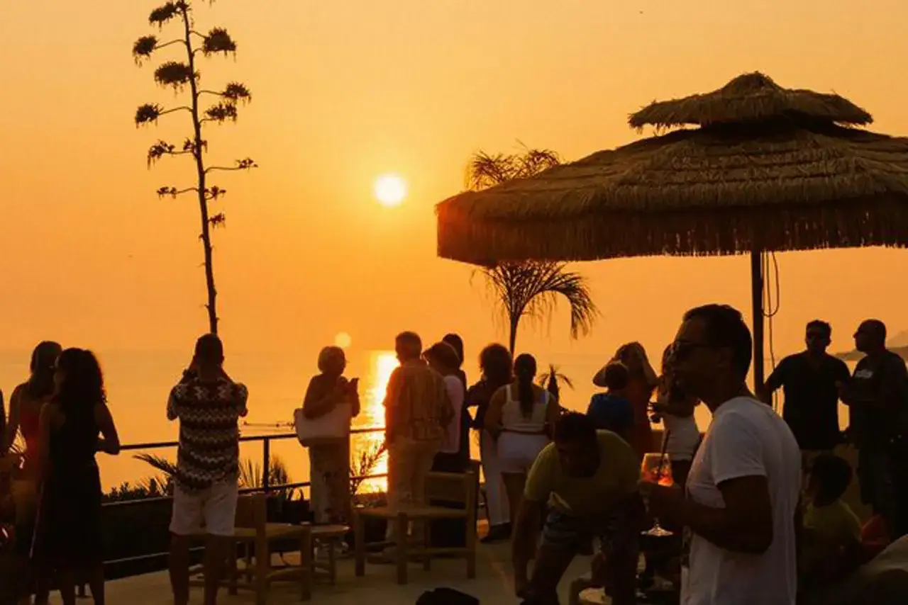 Persone godono di un tramonto dorato su una fi beach, con un grande ombrellone di paglia e una palma.