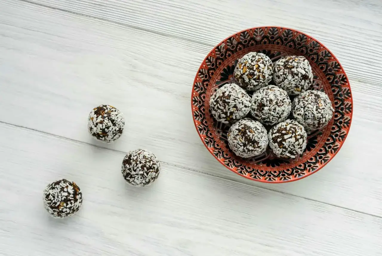 En sk&aring;l med nyttiga chokladbollar, rullade i kokosflingor, st&aring;r p&aring; ett vitt tr&auml;bord. N&aring;gra bollar ligger utspridda bredvid.