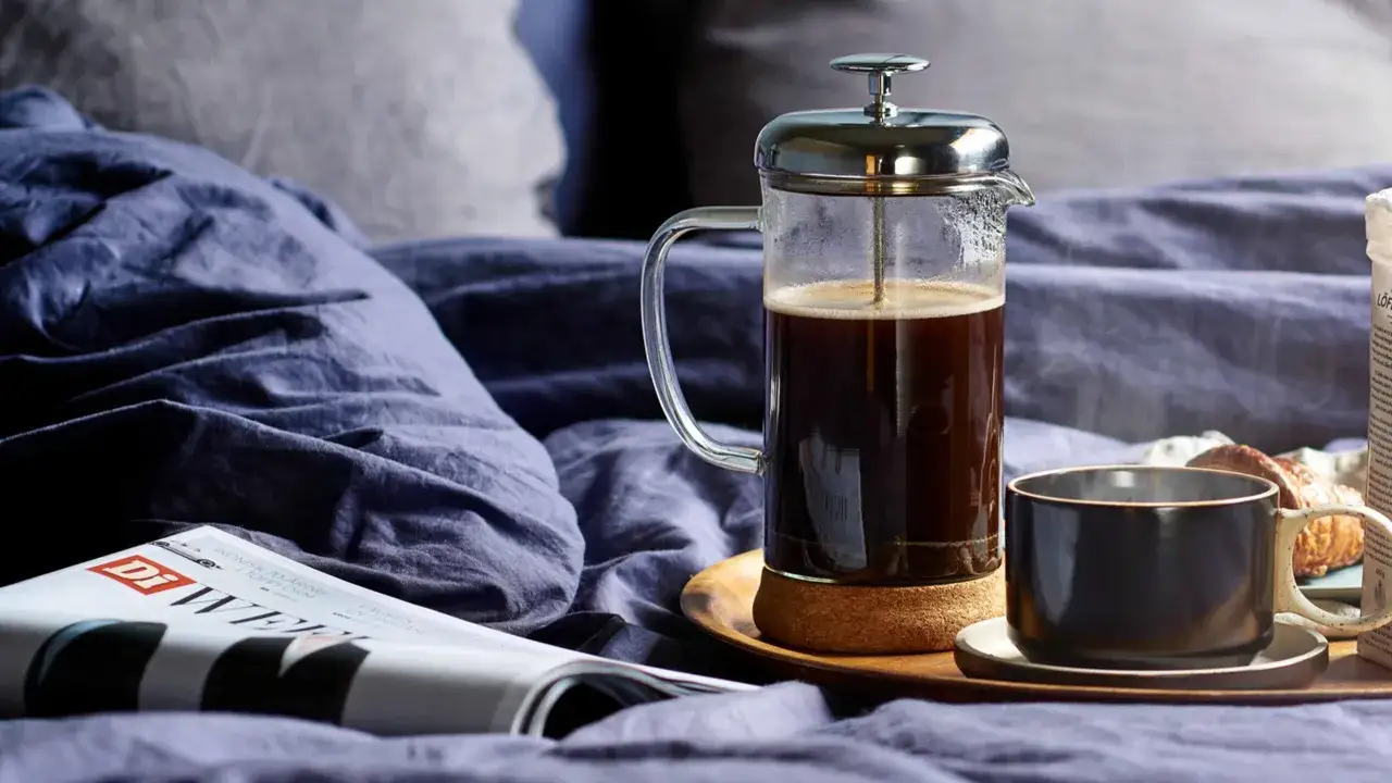 En franskpress med gott kaffe står på en bricka i sängen bredvid en tidning och en croissant. En perfekt morgon.