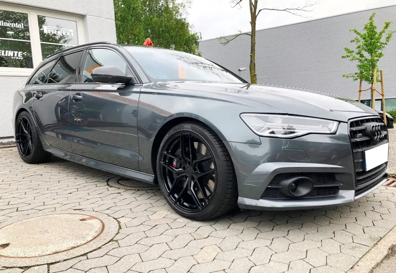 Szary Audi S6 C7 Avant na czarnych felgach, zaparkowany na kostce brukowej przed budynkiem z logo Pirelli.