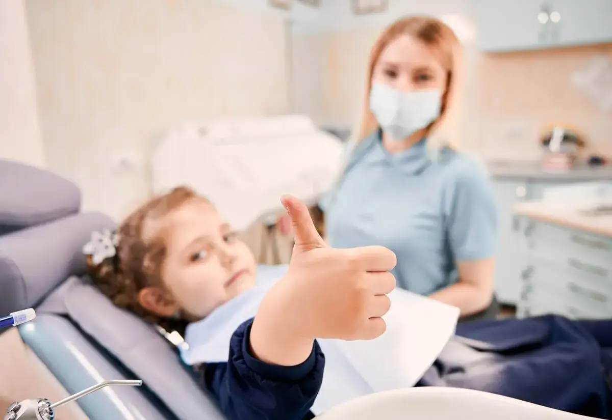 Uśmiechnięta dziewczynka po zabiegu lakowania zębów pokazuje kciuk w górę. Obok stoi dentystka w maseczce.