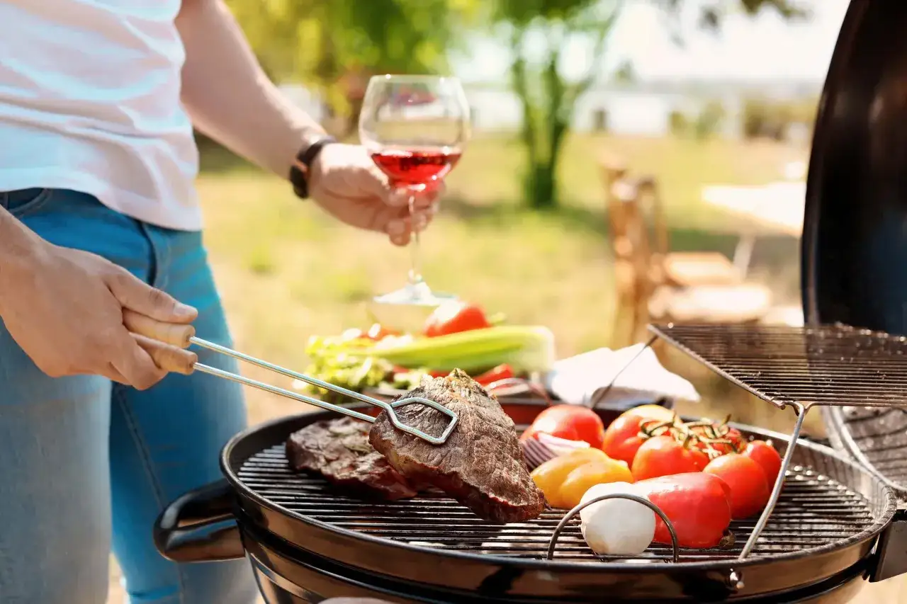 Un homme prépare une grillade avec des steaks et des légumes, un verre de vin rouge à la main. Parfait pour un repas en plein air.