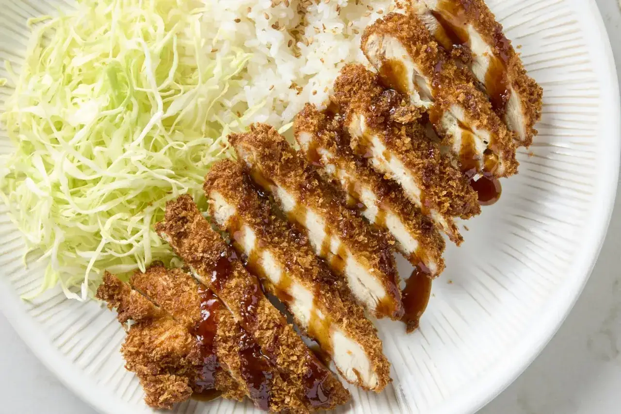 Krispigt kyckling katsu med ris och strimlad k&aring;l, allt serverat p&aring; en vit tallrik med en fin textur.