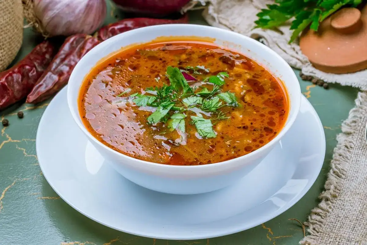 Aromaticzna zupa charczo, bogata w smaku, ozdobiona świeżą natką pietruszki i kopru. Obok suszone papryczki chili i cebula.