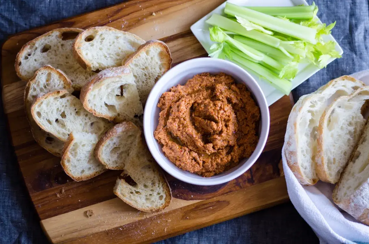 En skål med muhammara, perfekt för ett muhammara recept, serveras med skivat bröd och selleristavar på en träskärbräda.