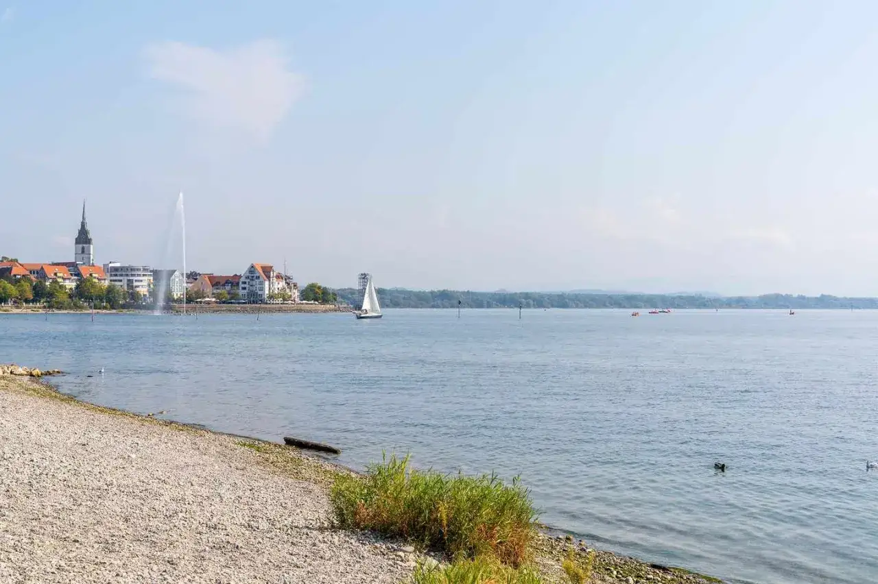 Friedrichshafen am Bodensee: Ein Segelboot gleitet &uuml;ber das Wasser, im Hintergrund die Stadt mit einem hohen Brunnen.