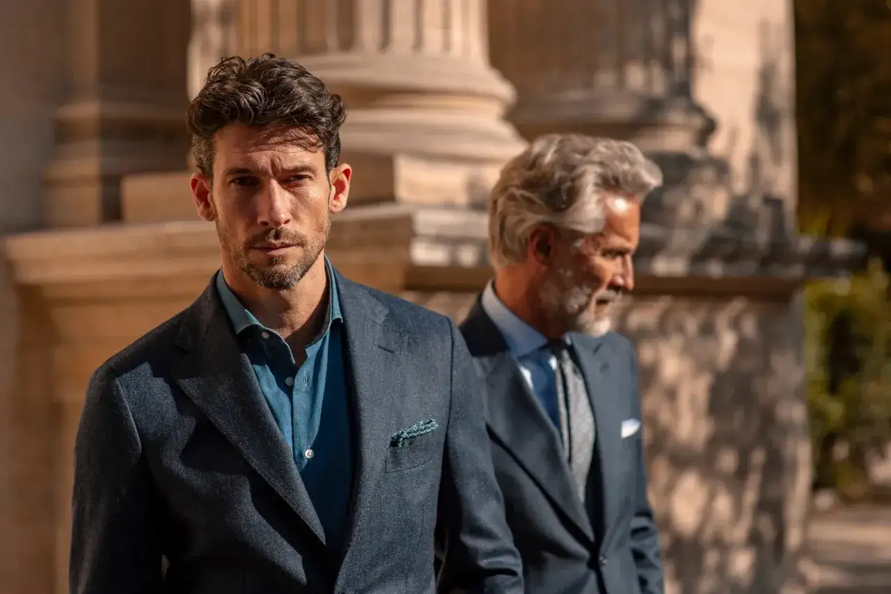 Due uomini eleganti in abito spezzato, uno in primo piano con giacca blu e camicia denim, l'altro sfocato sullo sfondo.