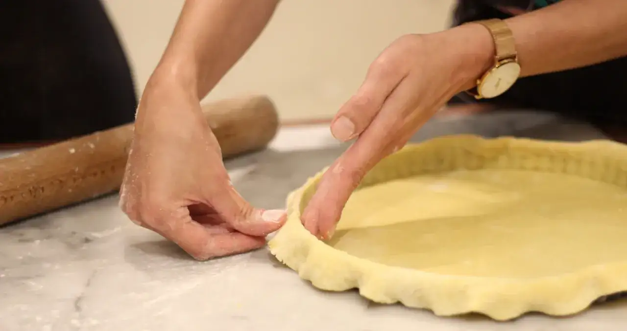 Mains façonnant la pâte à foncer dans un moule à tarte. Un rouleau à pâtisserie est visible à gauche.