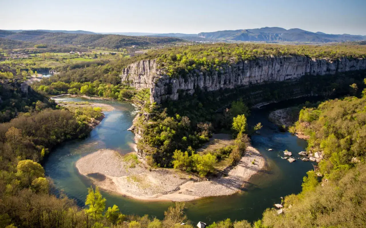 Vue aérienne d'une rivière sinueuse traversant un paysage verdoyant, bordée de falaises rocheuses. Un cirque de gens pourrait s'y retrouver pour admirer la beauté naturelle.