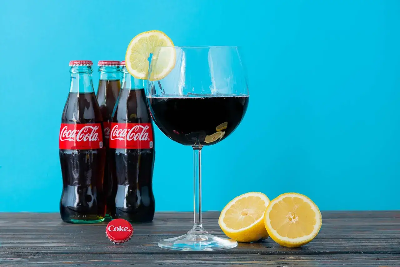 Kieliszek z ciemnym płynem, ozdobiony plasterkiem cytryny, obok butelek Coca-Coli i połówki cytryny. Idealny na letnie wieczory, ten drink to wino z colą.