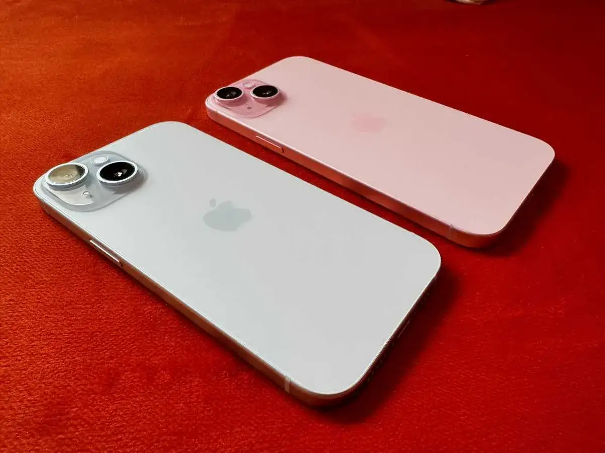 Två iPhones, en vit och en rosa, ligger på en röd yta. Den vita telefonen är större, vilket ger en uppfattning om iphone 15 pro max storlek.