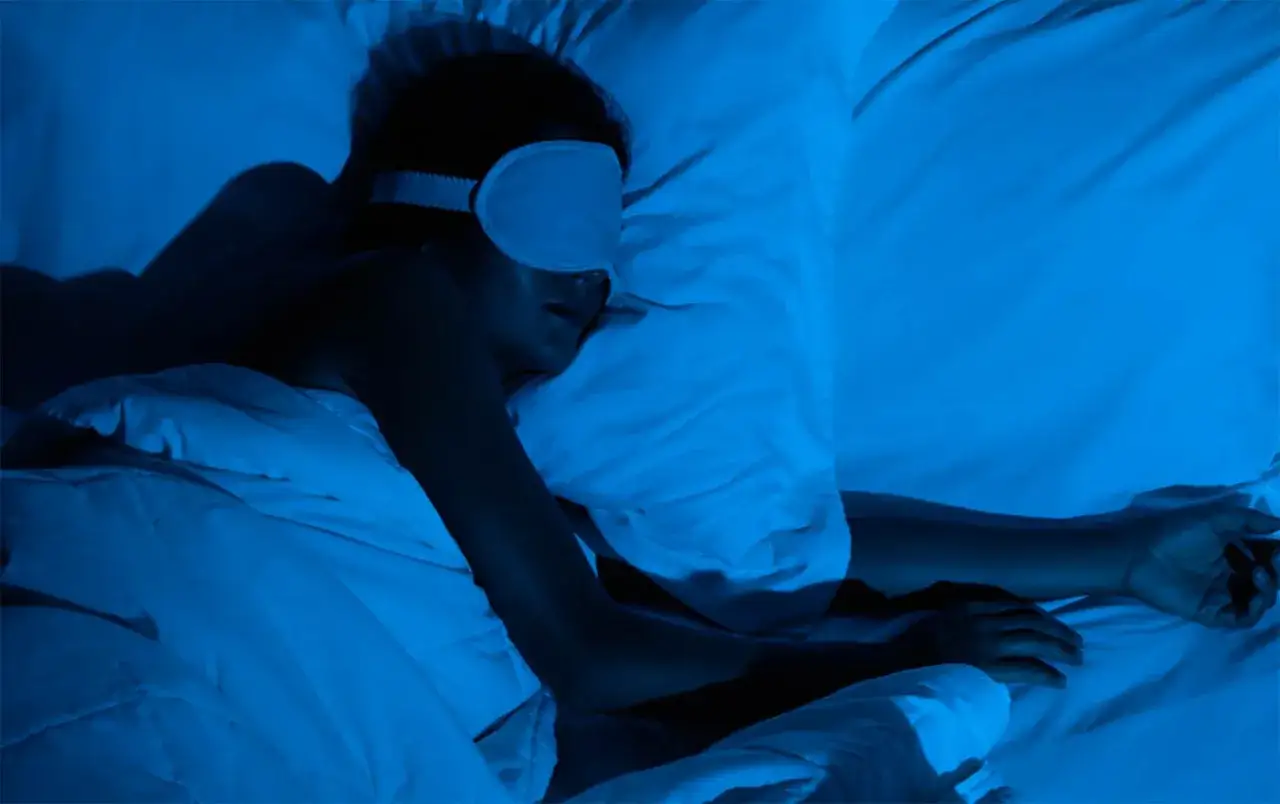 Persona duerme con antifaz, envuelta en mantas. Se siente como si me levanto cansado, incluso en la oscuridad azulada.