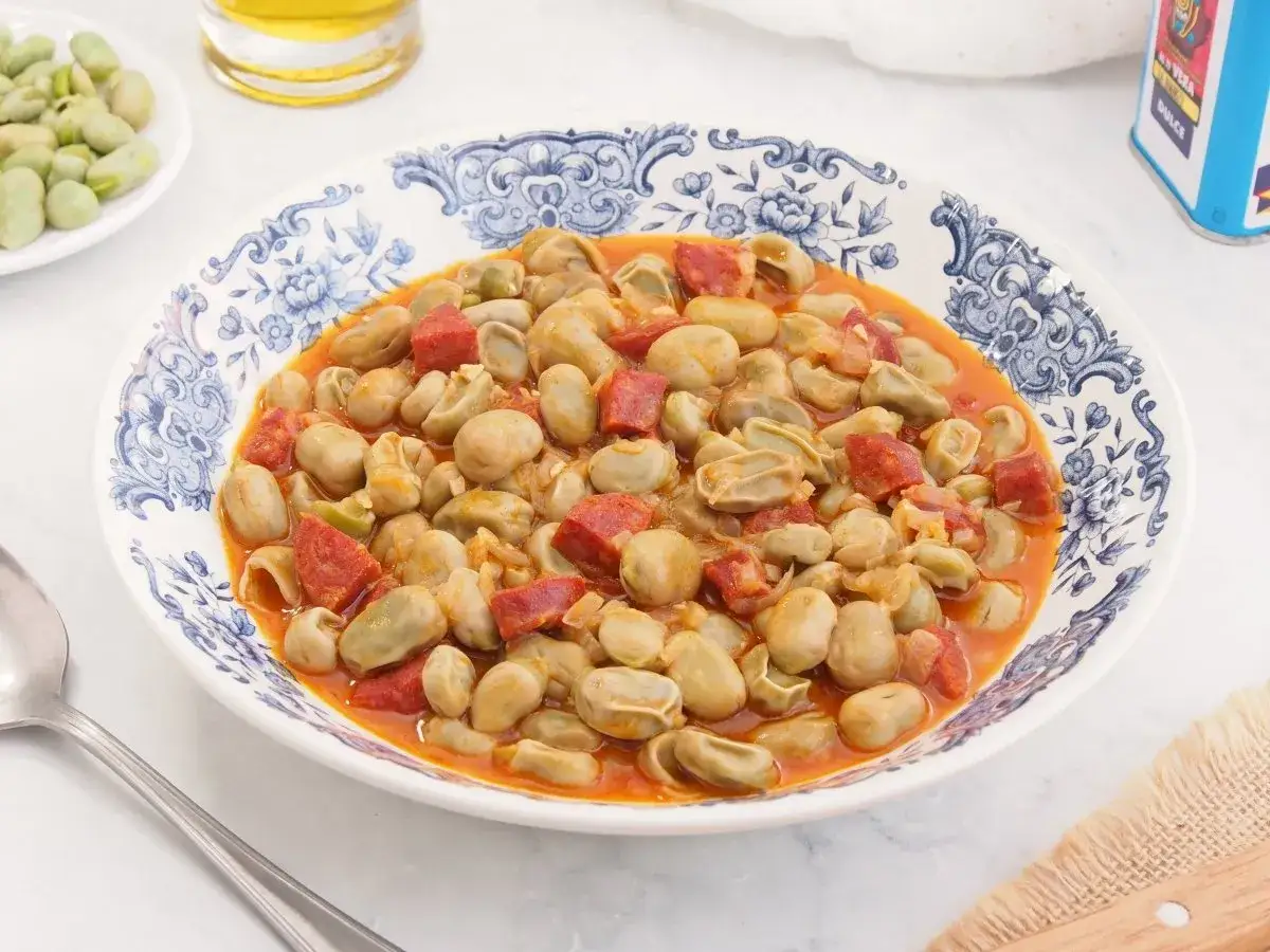 Plato de habas tiernas guisadas con trozos de chorizo español en una salsa rojiza, servido en una vajilla azul y blanca.
