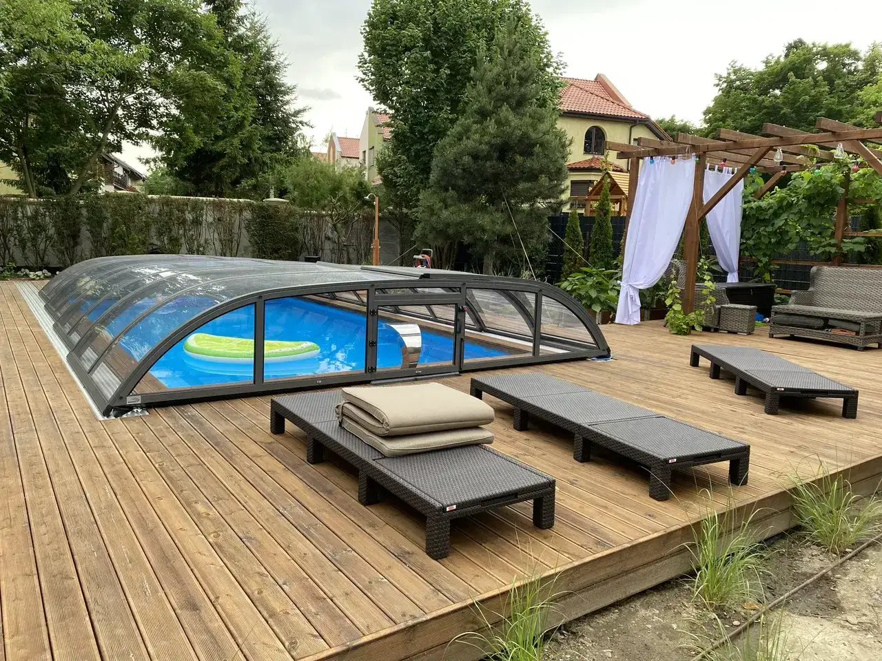 Zabudowany deskami basen z zieloną dmuchaną wyspą, otoczony leżakami i drewnianą pergolą.