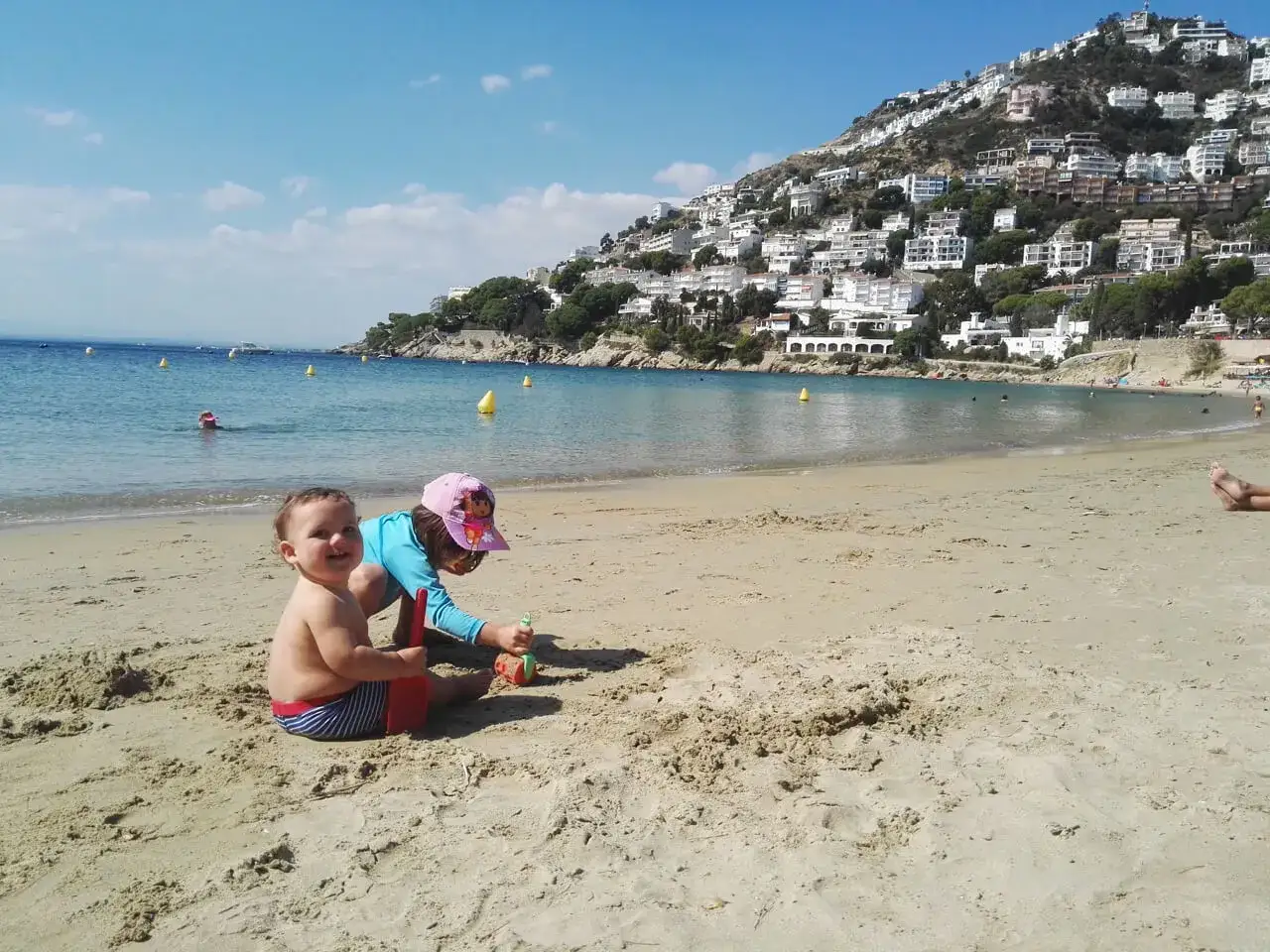 Dos niños juegan en la arena de una playa en Cataluña, ideal para saber donde ir con niños de 2 a 3 años. El mar y un pueblo en la colina completan la escena.