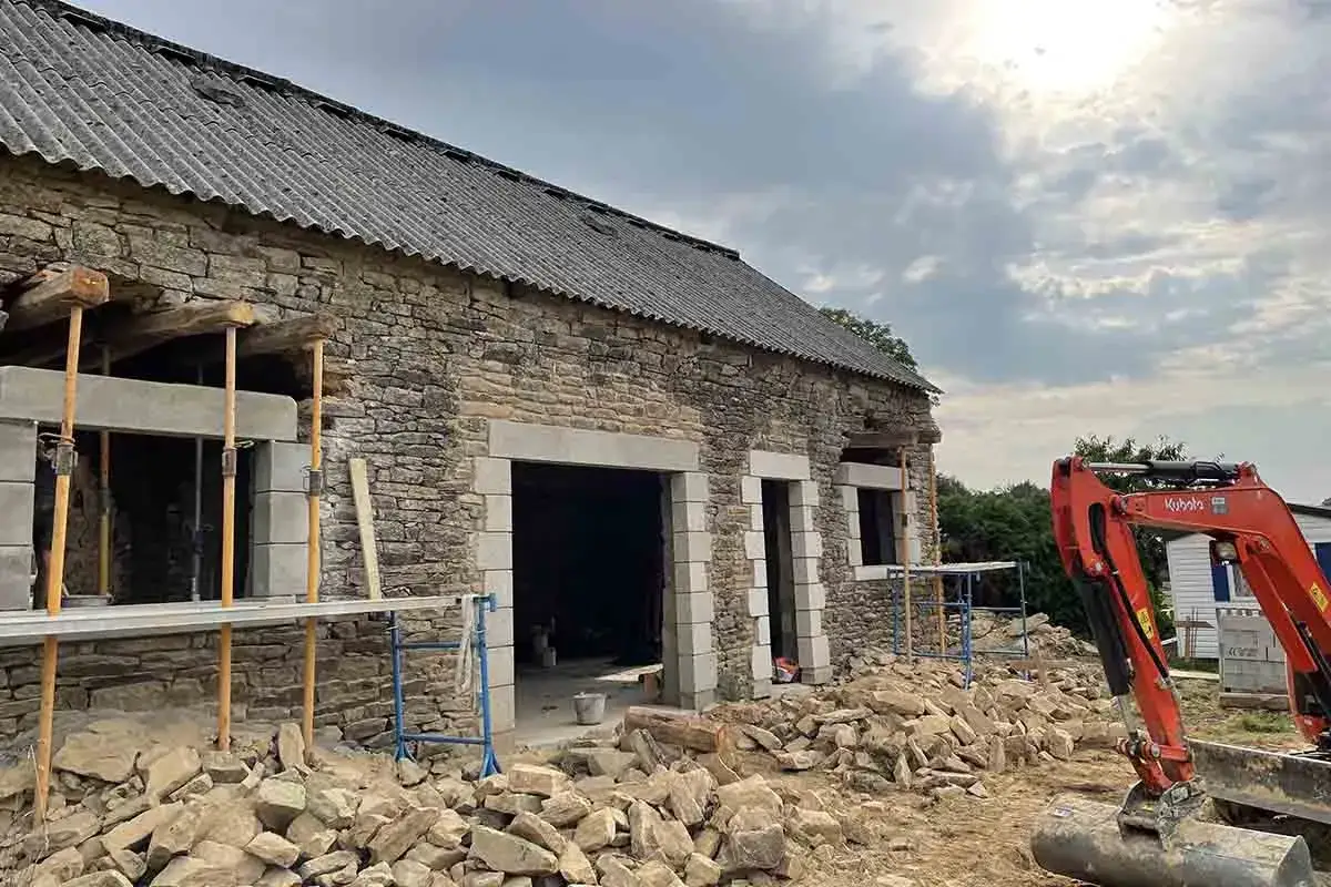 Chantier de maçonnerie : une maison en pierre avec une grande ouverture et une fenêtre en cours de finition. Des échafaudages et des pierres jonchent le sol.