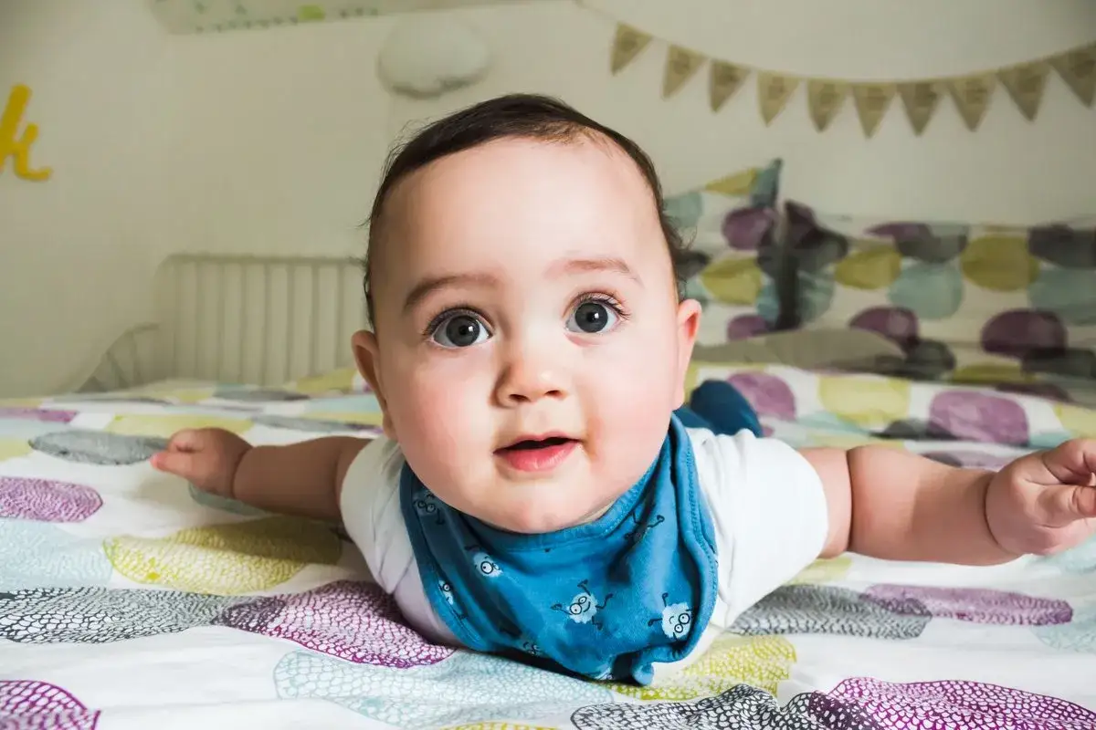 Un adorabile neonato di 4 mesi, sdraiato a pancia in giù sul letto, con gli occhi spalancati e un bavaglino blu.