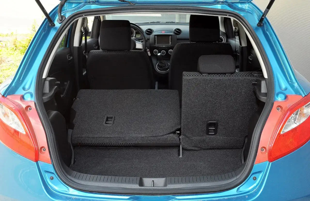 Błękitna Mazda 2 z 2008 roku z otwartym bagażnikiem. Tylne siedzenia złożone, tworząc dużą przestrzeń ładunkową.