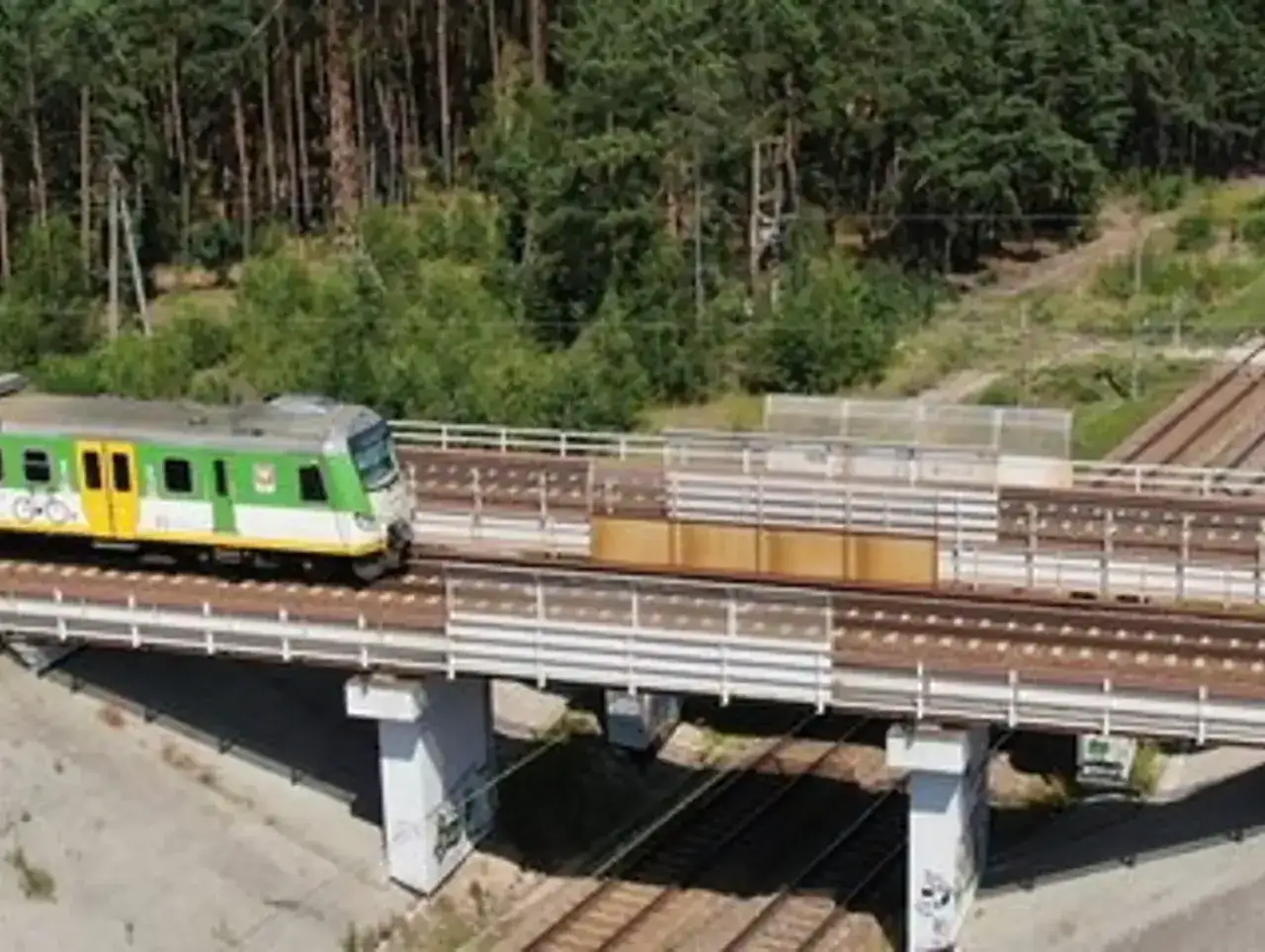 Zielono-ż&oacute;łty lekki pojazd szynowy przejeżdża przez wiadukt pośr&oacute;d lasu.