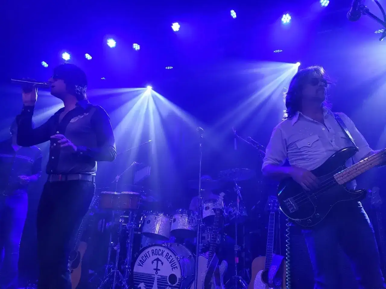 Zespół gra yacht rock, wokalista śpiewa, basista gra na gitarze basowej, w tle perkusja.