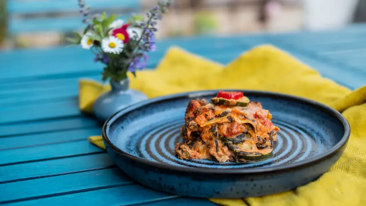 Ein St&uuml;ck k&ouml;stliche vegetarische Lasagne mit Zucchini und Tomaten auf einem blauen Teller, daneben ein kleiner Blumenstrau&szlig;.