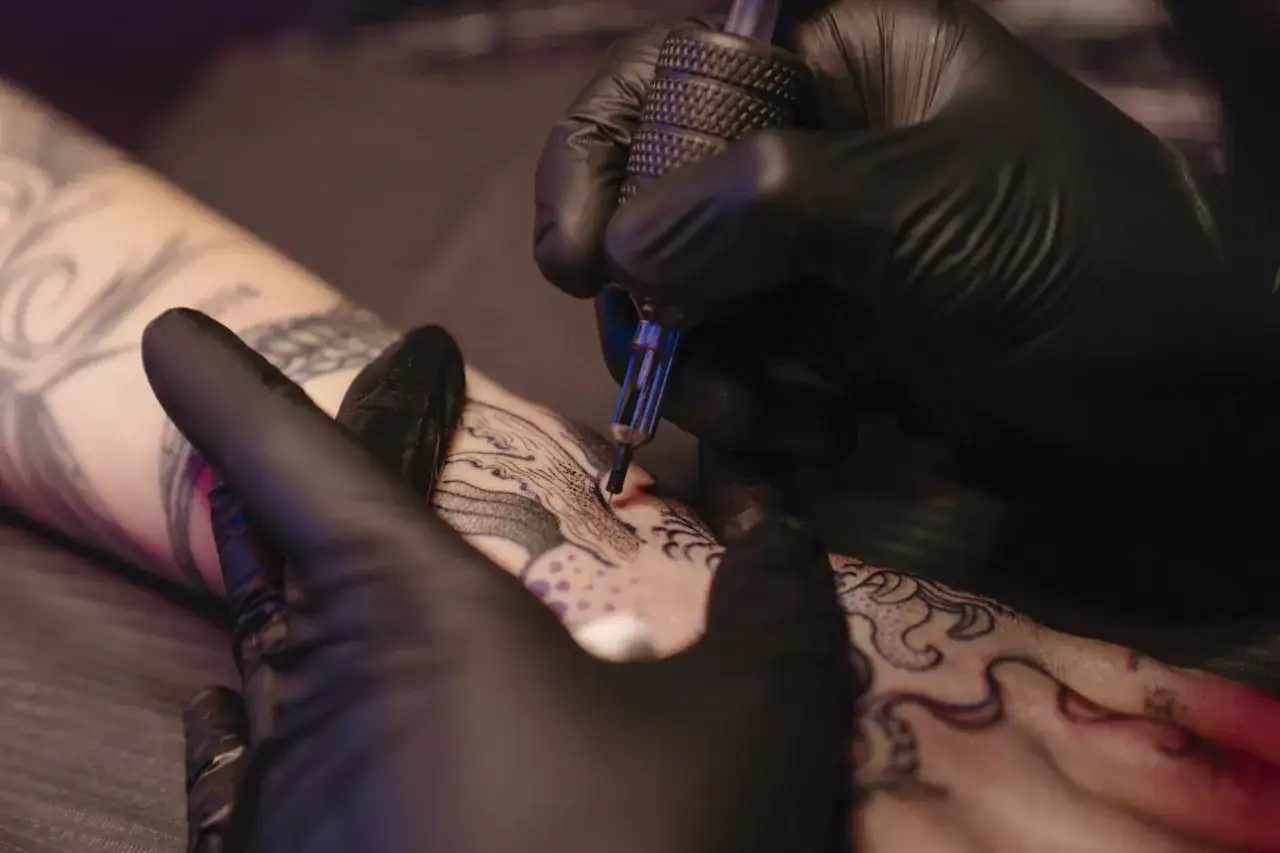 Manos enguantadas trabajando en un tatuaje intrincado. El artista usa una máquina de tatuar para aplicar **buena tinta tattoo** en la piel, creando un diseño detallado.