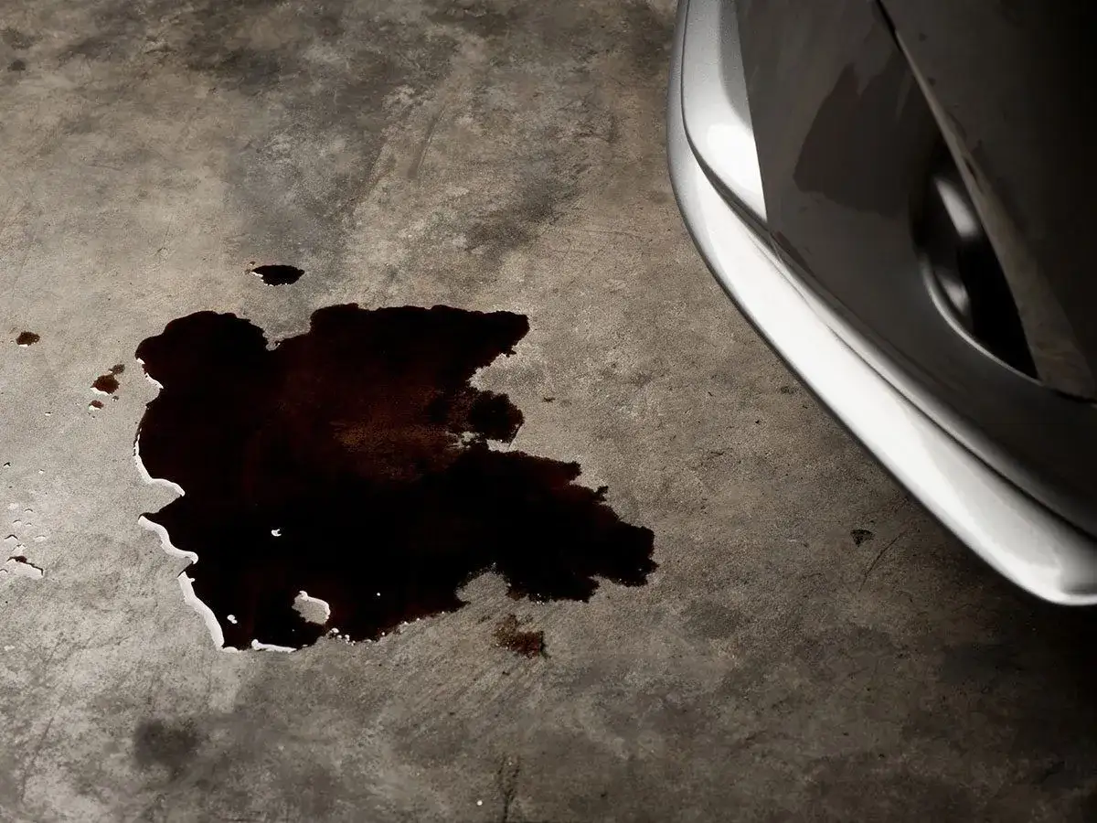 Mancha oscura de aceite en el suelo de hormigón, indicando una **pérdida de aceite por el cárter** de un coche plateado.