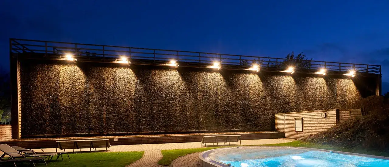 Abendliche Szene an der **Therme Sauerland**: Ein gro&szlig;es Gradierwerk mit beleuchteten Reisigw&auml;nden, davor ein Pool und Sitzb&auml;nke.