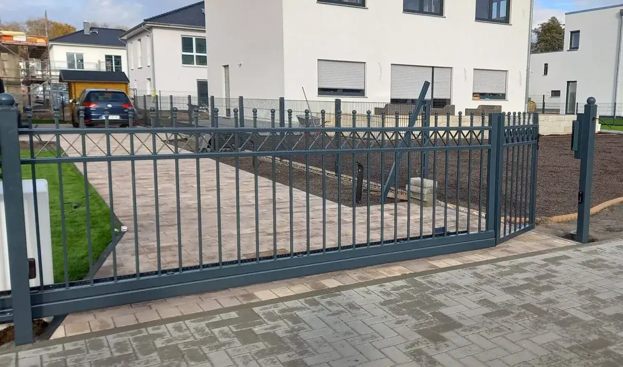 Nowa, metalowa brama z furtką bez słupka, z ozdobnymi kulkami na górze, prowadzi do nowoczesnego domu.