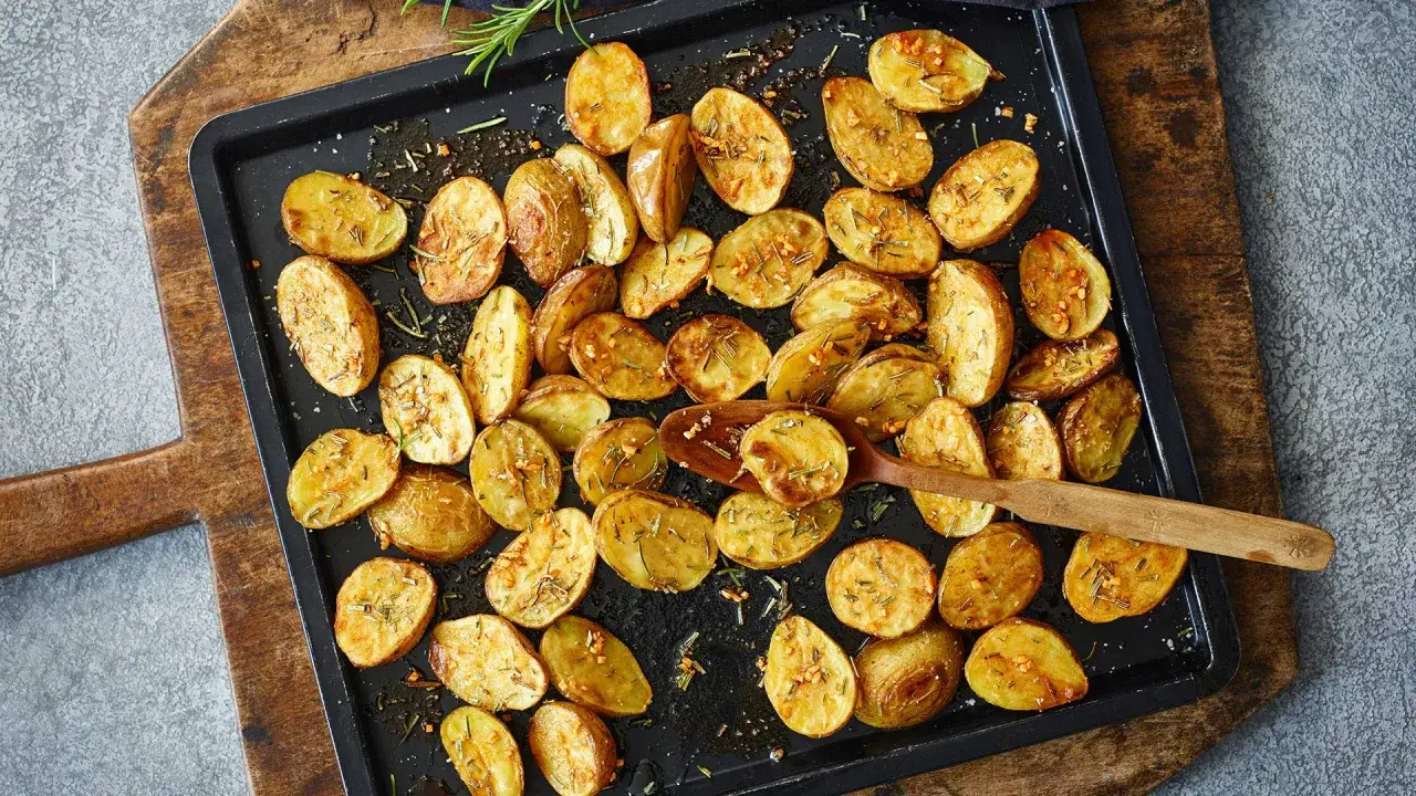 Goldbraune, knusprige Rosmarinkartoffeln auf einem Backblech, mit frischem Rosmarin und Knoblauch verfeinert. Ein Holzl&ouml;ffel liegt auf den Kartoffeln.