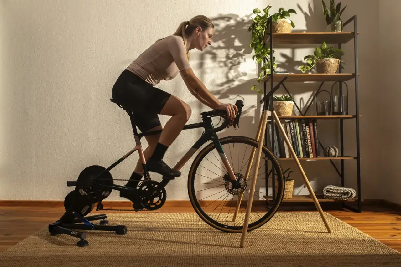 Kobieta trenuje na rowerze stacjonarnym, korzystając z aplikacji do trenażera. W tle regał z roślinami i książkami.