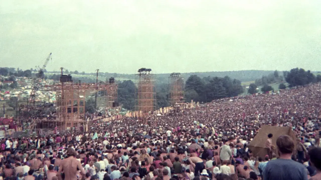 Tłum ludzi na festiwalu, gdzie jest Woodstock. Widoczna scena i wieże głośnikowe.