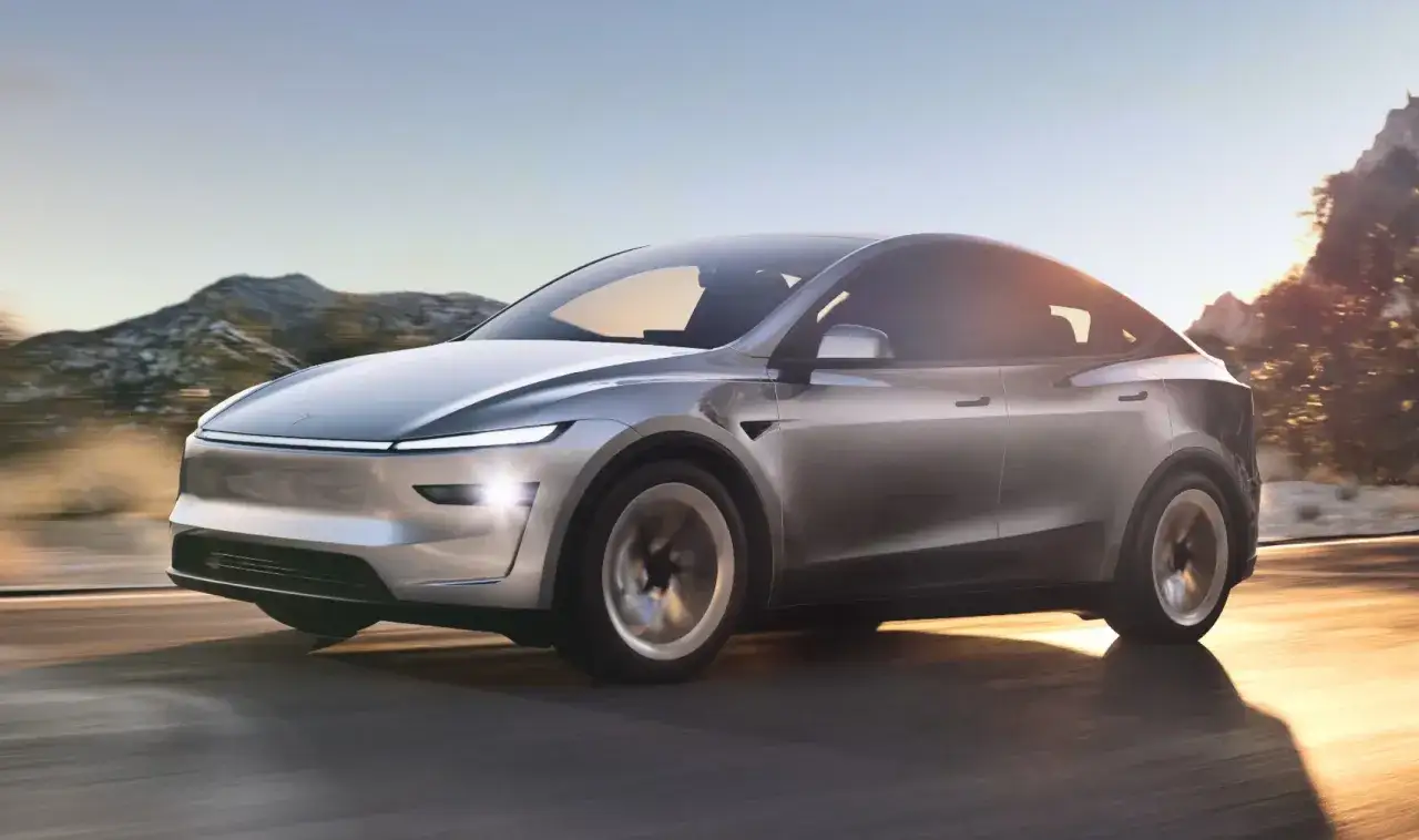 Srebrna Tesla Model Y pędzi po drodze, gdy zapada zmierzch. G&oacute;ry i drzewa w tle tworzą malowniczy krajobraz.