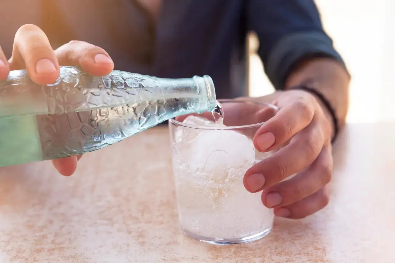 Une main verse de l'eau gazeuse dans un verre rempli de glaçons. Une boisson rafraîchissante pour se désaltérer.