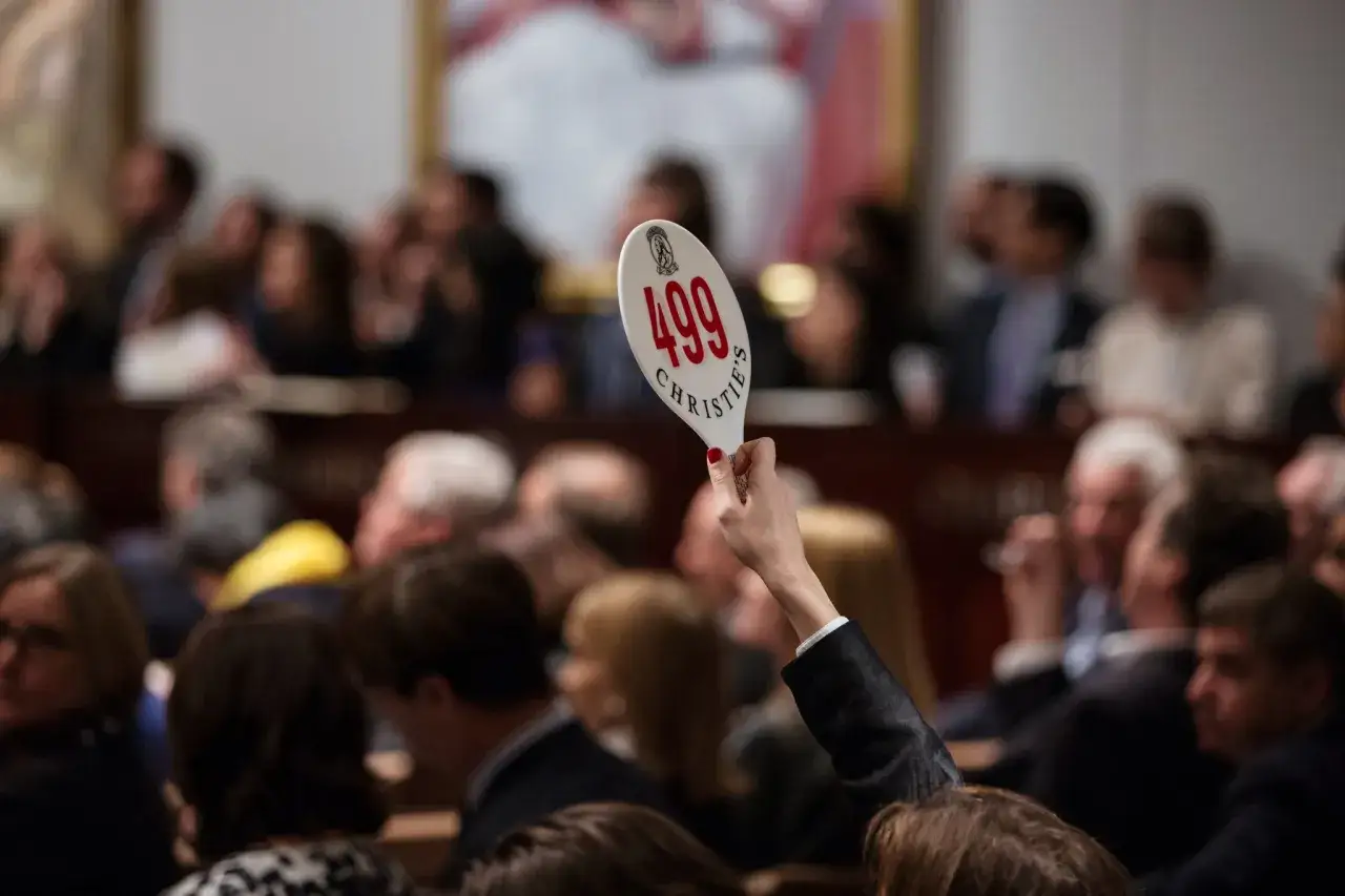 A hand holds up paddle #499 at a Christie's auction, with blurred attendees in the background. This scene evokes the high stakes of art sales, similar to those at Sotheby's.