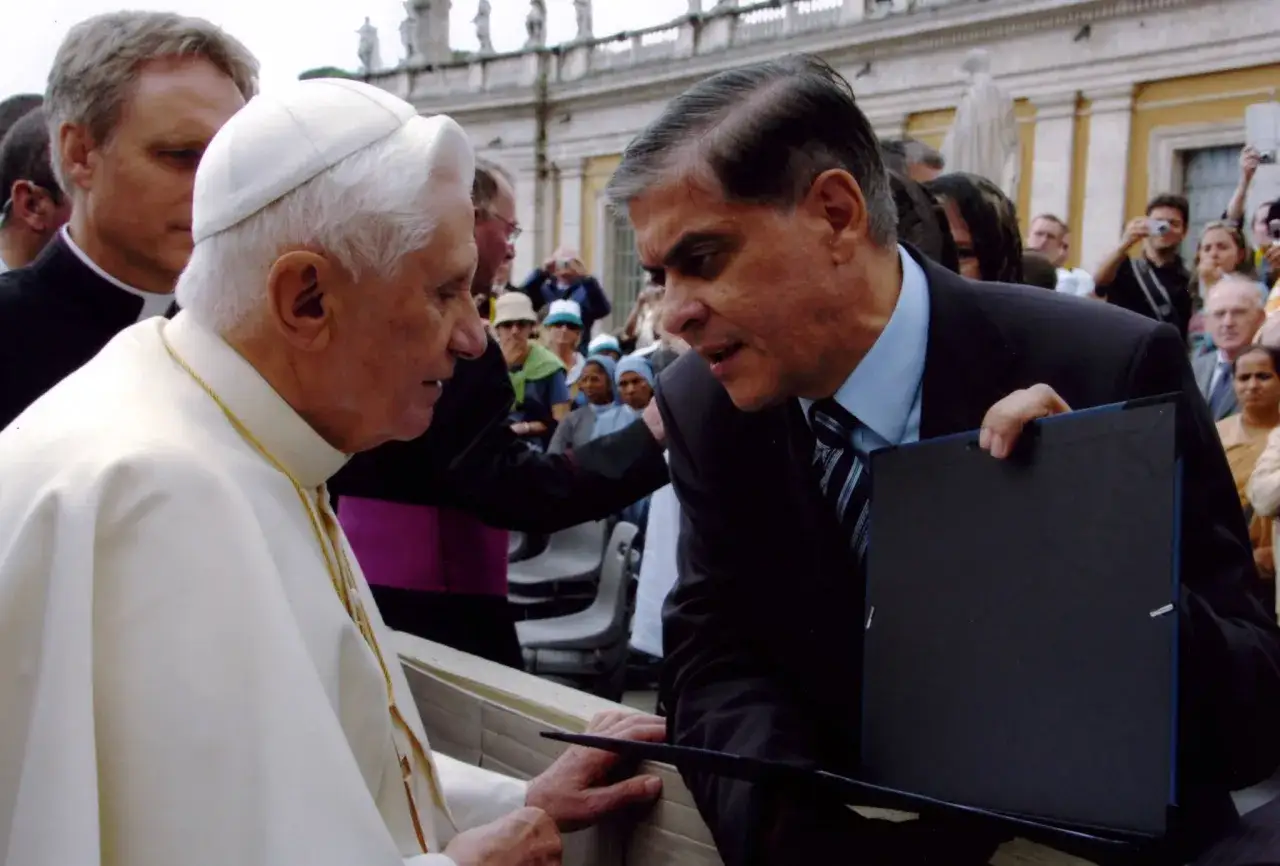 Benedikt XVI. im Gespr&auml;ch mit einem Mann, der ein schwarzes Mappe h&auml;lt. Im Hintergrund stehen Menschen.