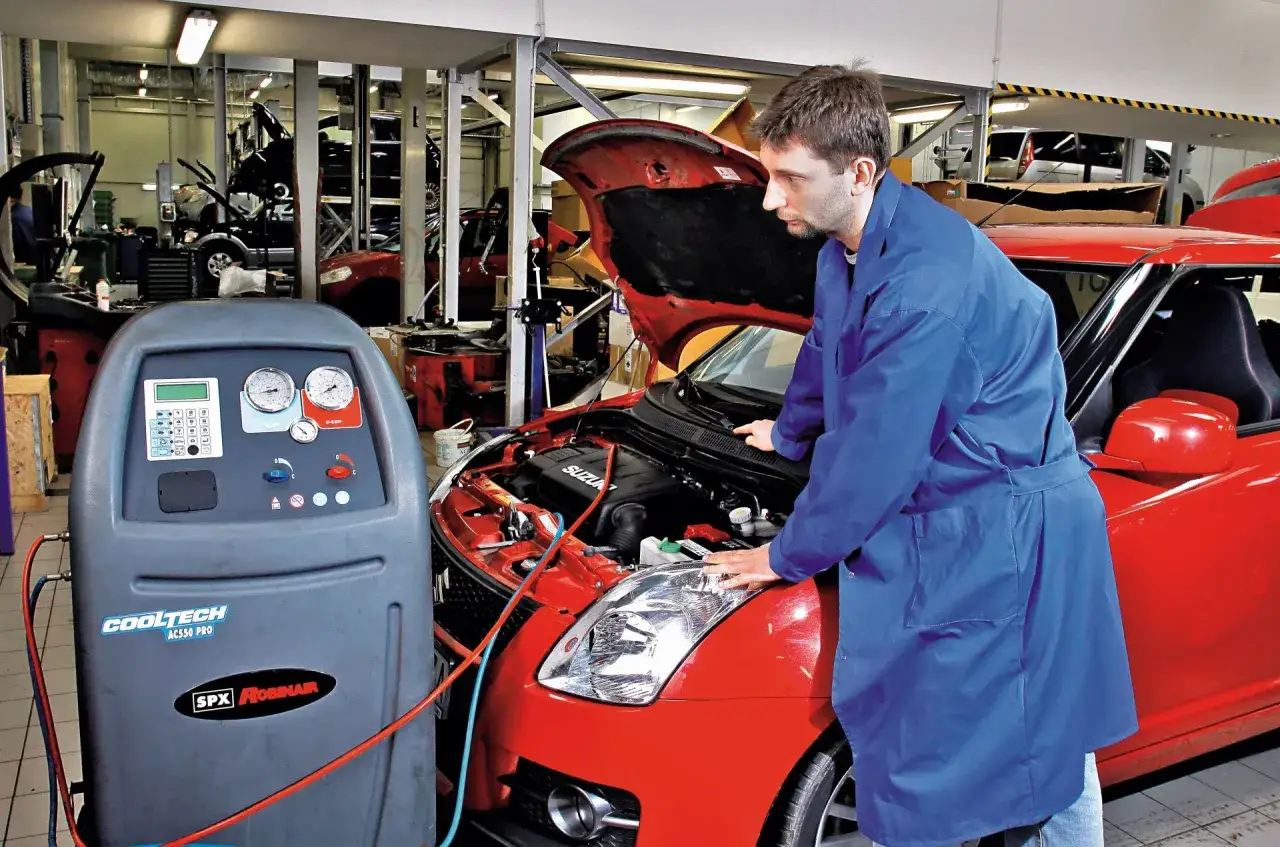 Mechanik w niebieskim fartuchu sprawdza działanie klimatyzacji w czerwonym samochodzie. Dowiedz się, ile kosztuje nabicie klimatyzacji.