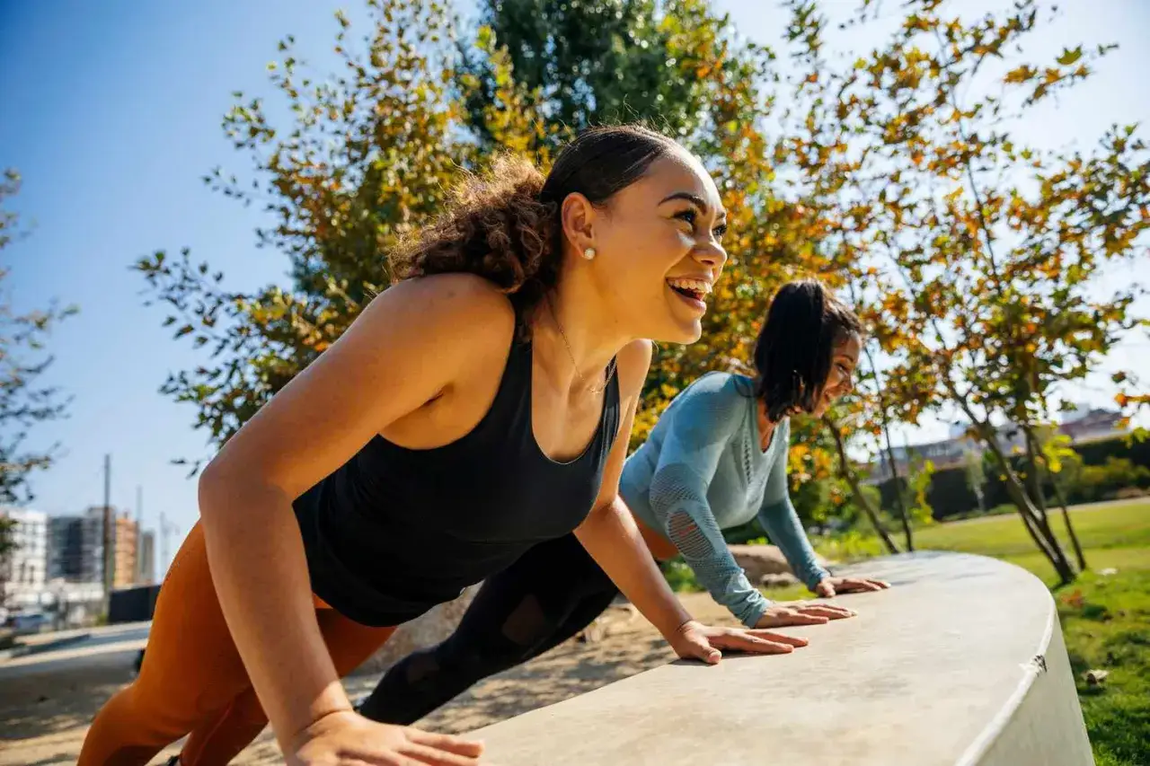 Zwei Frauen machen Liegest&uuml;tze im Freien. Sie lachen und genie&szlig;en ihr liss cardio Training unter B&auml;umen.