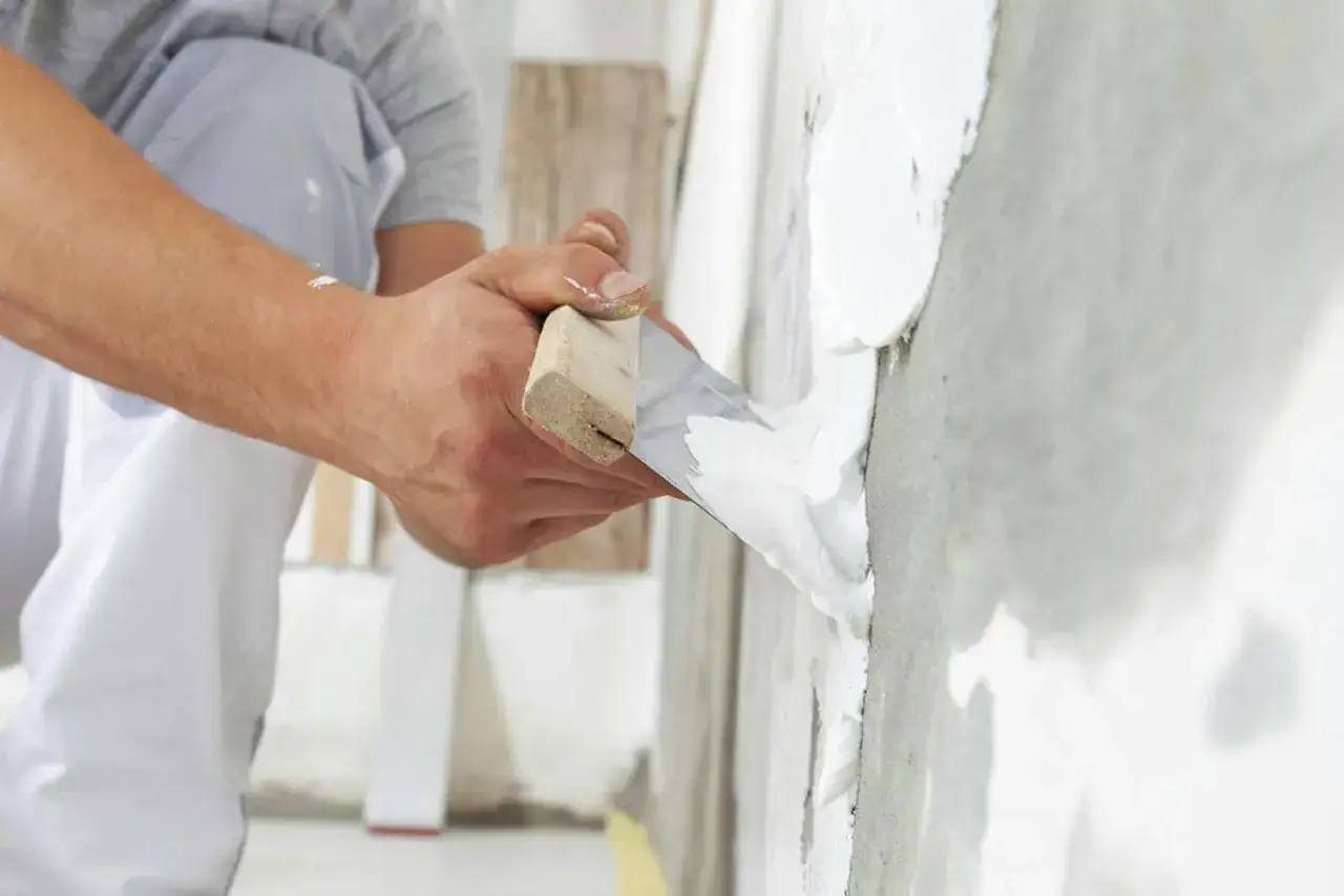 H&auml;nder med spackel och spackelspade applicerar putsbruk inomhus p&aring; en v&auml;gg.