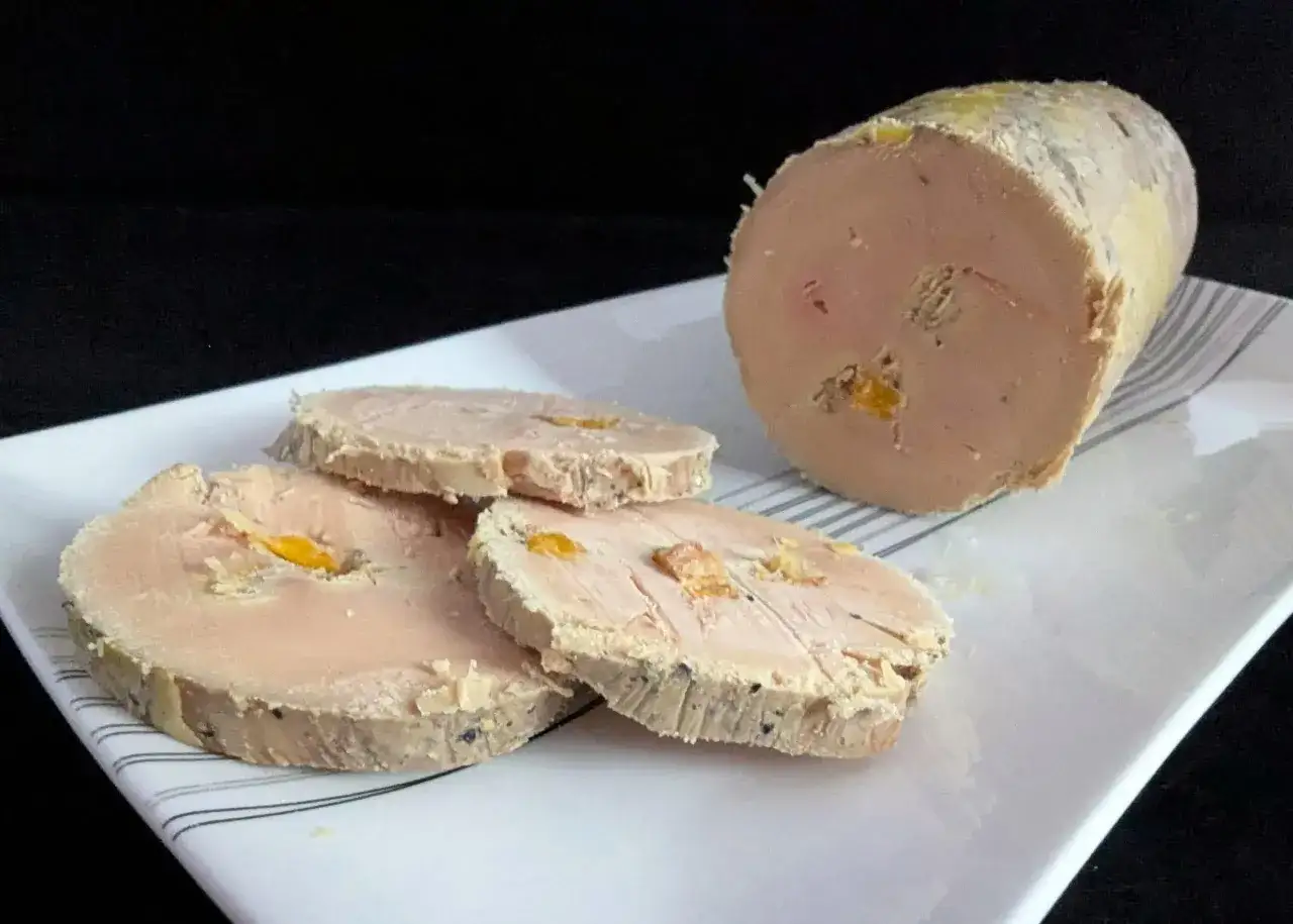 Tranche de foie gras maison, parsemée de morceaux orangés, sur une assiette blanche rayée. Un délice artisanal.