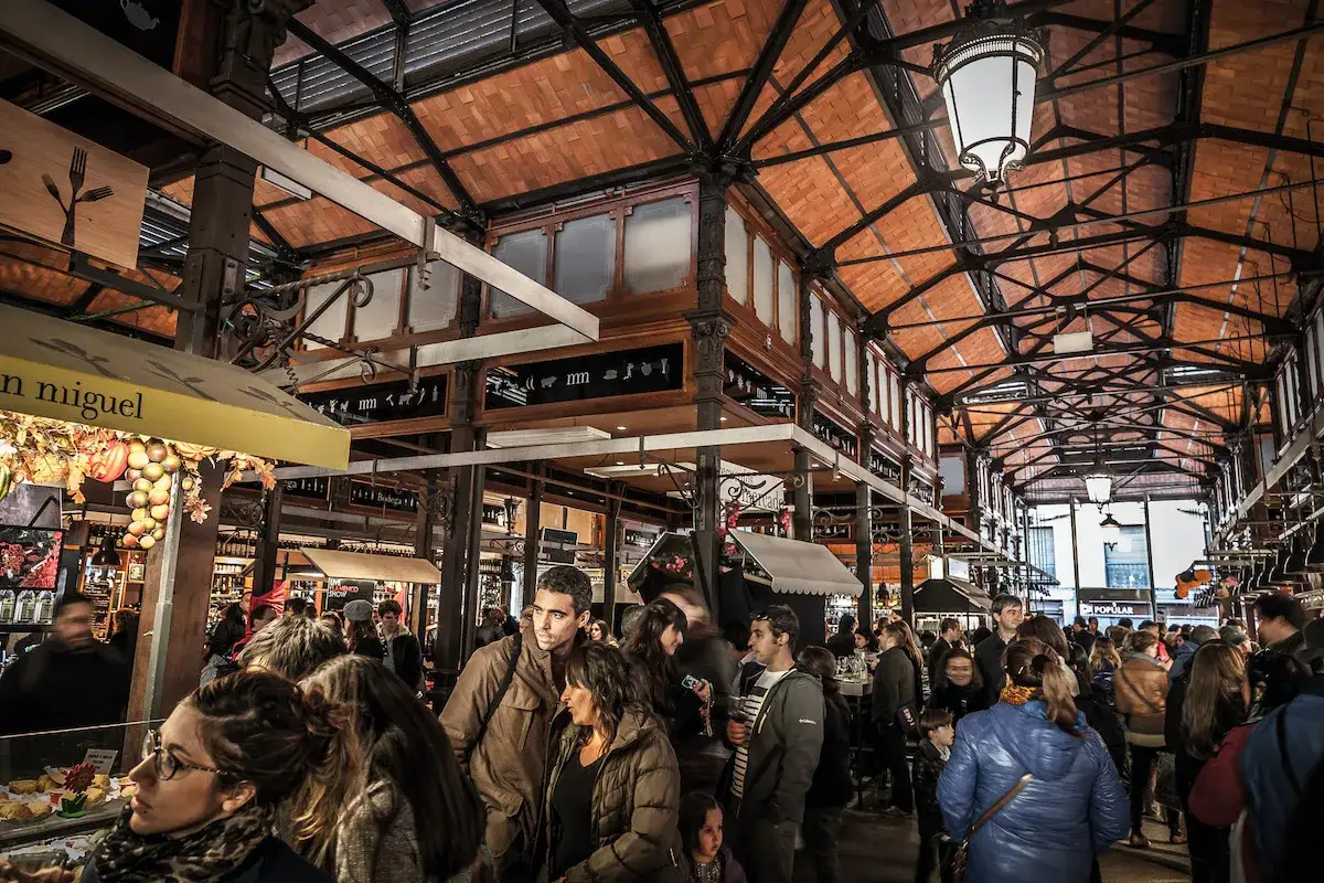 Menschenmassen schlendern durch den belebten Mercado de San Miguel unter einem beeindruckenden Ziegeldach.