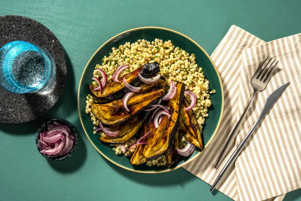 Deliciosas recetas con berenjenas: berenjenas asadas con cuscús y cebolla morada, acompañadas de agua con gas.